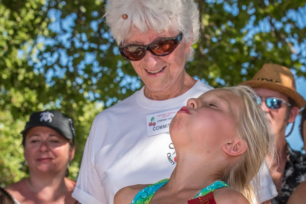 While volunteer Jessie Smythe looks on, a young girl fires a cherry pit into the air during the cherry pit spitting contest at Cherry Fiesta. (Richard McGuire photo)