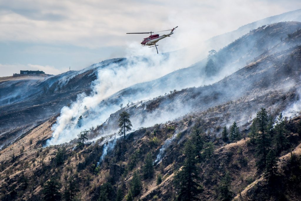 A helicopter flies over burning hills as it carries water from a slough to the Testalinden Creek fire near Road 20 and Highway 97, just after noon Saturday. (Richard McGuire photo)