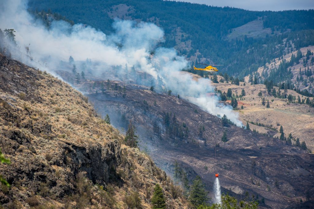 A helicopter buckets water onto fire burning down the Testalinden Creek Valley after picking it up at a nearby slough. This photo was taken at 12:10 p.m. Saturday.