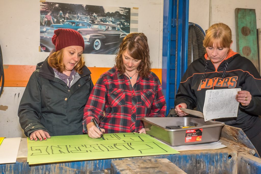 Sharlene Fernandes (left) and Patricia Pearson worked on signs in an effort to keep schools open while Brenda Dorosz (right) reviews some notes. Dorosz chairs the Save Our Schools (SOS) committee, which plans to show a presence at a public consultation Tuesday night at 7 p.m. at Osoyoos Secondary School. The signmaking activities were underway Monday evening at JF Kustoms garage, which owner J.F. Launier made available. (Richard McGuire photo)