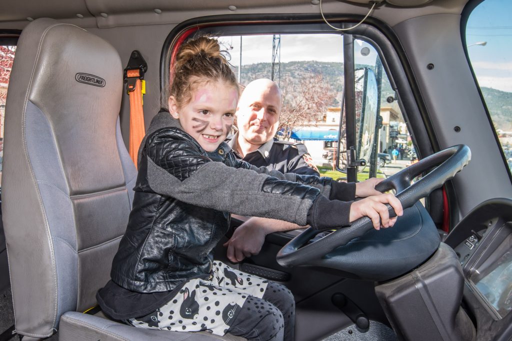 Brooke Young, 7, of Osoyoos imagines she's driving the fire truck as Osoyoos firefighter Casey Brouwer keeps an eye on her. In past years a few children have played with the controls, perhaps in an attempt to drive off with the truck, so this year only a few lucky ones got to sit in the driver's seat under close supervision. (Richard McGuire photo)