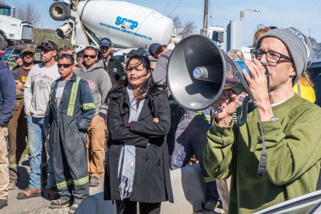 Jamie Elder of Unity Clothing addresses the crowd with the megaphone. In the background is a cement truck from South Okanagan Concrete Products, one of many trucks parked at Osoyoos Secondary School. (Richard McGuire photo)