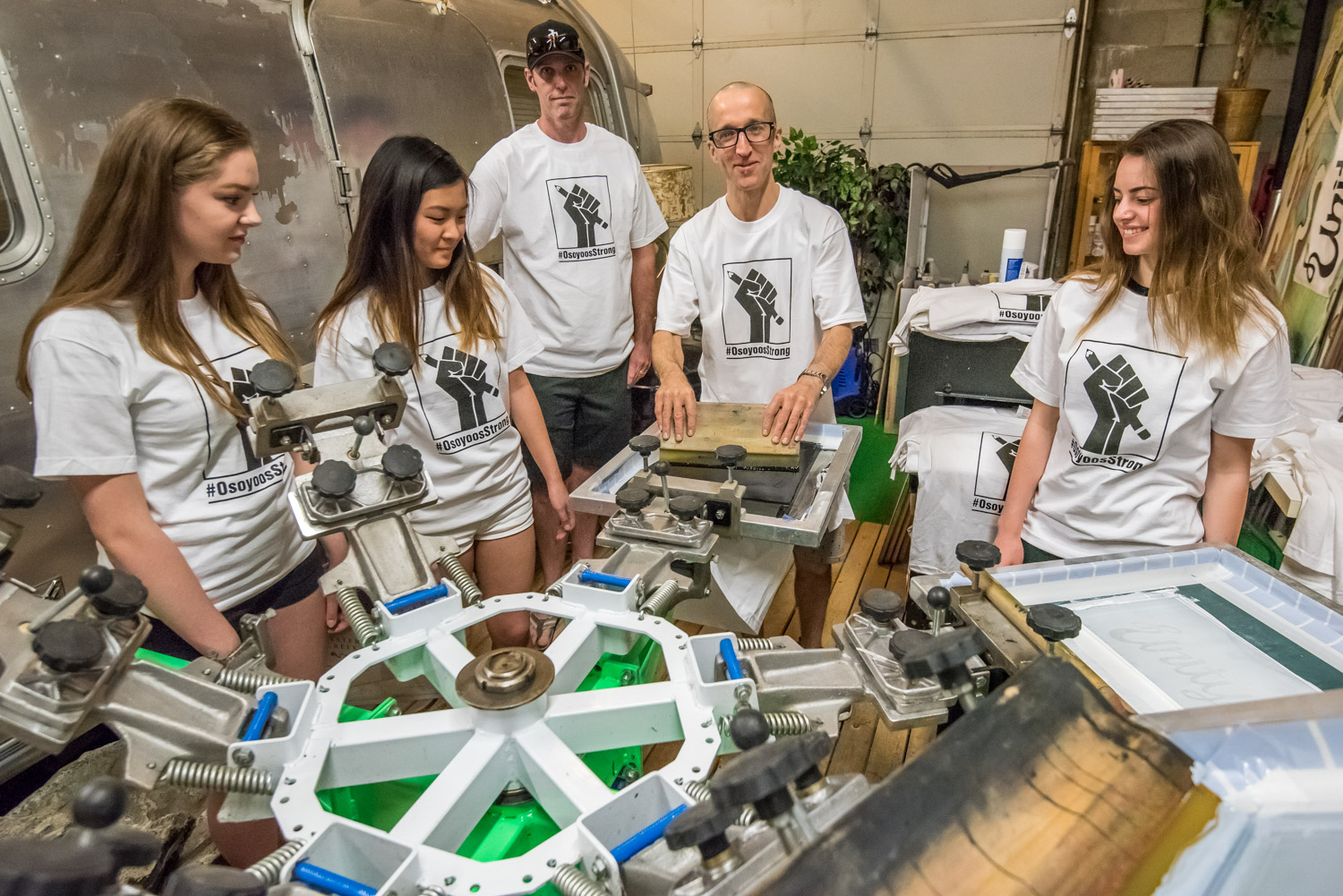 Jamie Elder (fourth from left), owner of Unity Clothing and Boardsports, prepares to silk screen t-shirts with the hashtag #OsoyoosStrong. Looking in from left are volunteers Hannah Shiels, Kelly Lao, Brock Jackson, Elder and Alyssa Macor. (Richard McGuire photo)