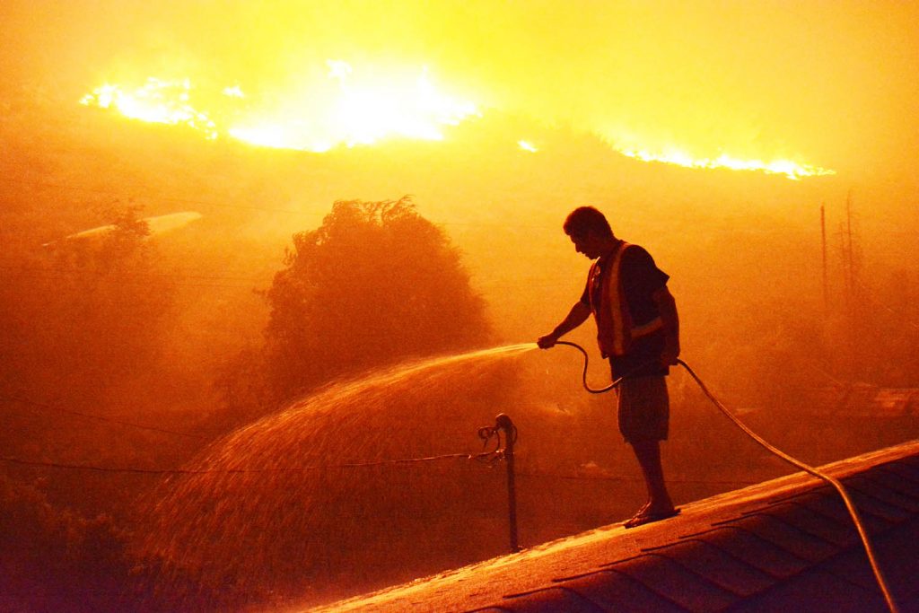 Spartan Street resident J.C. Oliveira from Oliver quickly hoses down his roof as the flames creep towards his backyard last Friday. Oliveira and many other families were evacuated that evening, but were allowed back the following day when the threat subsided. (Lyonel Doherty photo)
