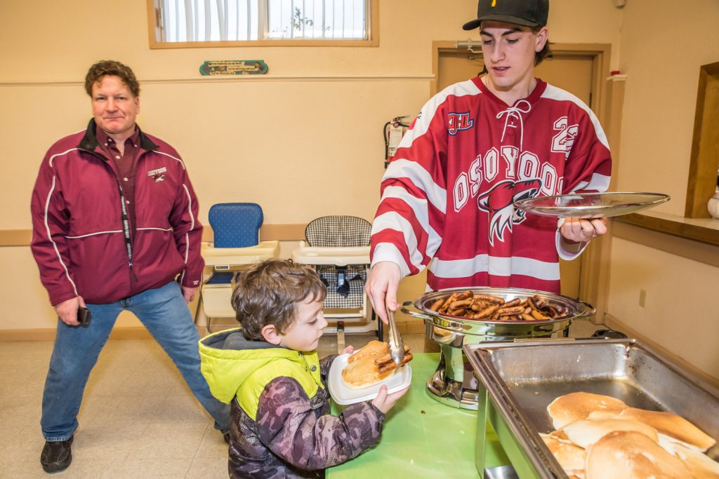 Coyote Ryan Morrell serves sausages to Elijah Zandvliet, of Osoyoos, who turns five this Saturday. At left is Coyotes owner Randy Bedard. The Coyotes were helping out at Breakfast with Santa and Photos at Osoyoos Baptist Church on Saturday morning. (Richard McGuire photo)