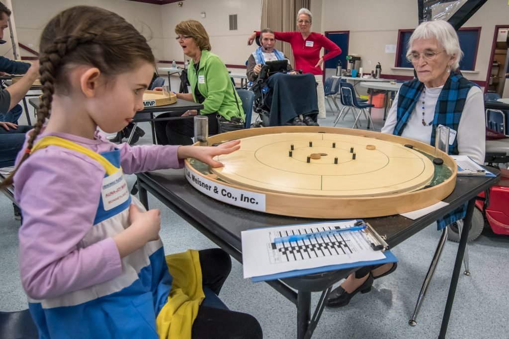 There was a 78-year age difference between them, but Pat Scott (right) of Oliver and Auna-Joy Mahan, formerly of Oliver and now from Bonney Lake, WA, enjoyed a round of crokinole together on Saturday. At 83, Scott was the oldest participant in the seventh annual B.C. Crokinole Championship and at five, Mahan was the youngest. Players from around the province and beyond participated in the event at the Oliver Community Centre. In the background in a wheelchair is event founder Clif Antypowich with his wife Joyce. Auna-Joy is their granddaughter. (Richard McGuire photo)