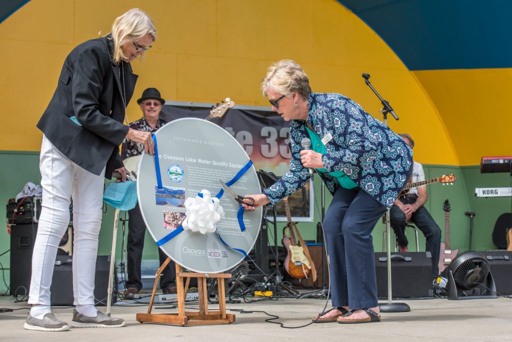 As Birgit Arnstein, president of the Osoyoos Lake Water Quality Society, steadies a new plaque, Mayor Sue McKortoff cuts the ribbon. The plaque will be installed by the walkway north of the museum. (Richard McGuire photo)