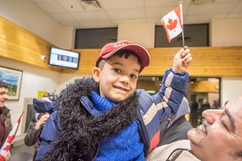 Hoisted in the air, Samer Tabanjat, 6, Osoyoos’ newest resident, waves a newly acquired Canadian flag and wears a ball cap with “Canada” on it as he arrives at Penticton Airport close to midnight Friday. The boy, his parents and older sister are Syrian refugees sponsored by a community fundraising campaign to settle in Osoyoos. He is held by Ayman Kanaan, who previously came to Summerland from Syria. (Richard McGuire photo)