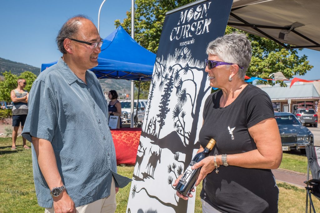 John Yap (left), the parliamentary secretary to B.C. Justice Minister Suzanne Anton, discusses wine with Beverly Forbes of Moon Curser Vineyards. Yap has been responsible for reforming B.C.'s liquor laws, including the change that allows local producers of beer, wine and spirits to sell and sample their products at farmers' markets. He toured Market on Main in Osoyoos on Saturday to find out how the program is working for local winemakers. (Richard McGuire photo)
